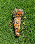 A single handmade Halimuyak smudge stick on green grass, showing intricate binding with dried marigolds and Almaciga resin.
